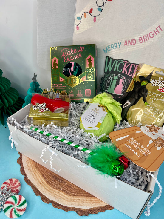 A gift box with a 'Wicked' theme containing various beauty products and a decorative ornament, wrapped in white paper with green and red accents, and a 'Happy Holidays' sign in the background.