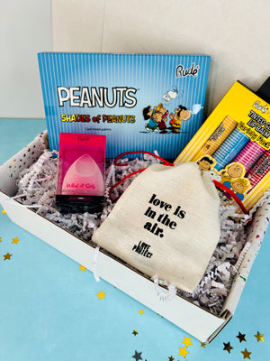 A gift box with ribbon, containing various beauty products including lip balm, eyeshadow, a bracelet, and a beauty sponge, with 'Peanuts' branding visible on the packaging.