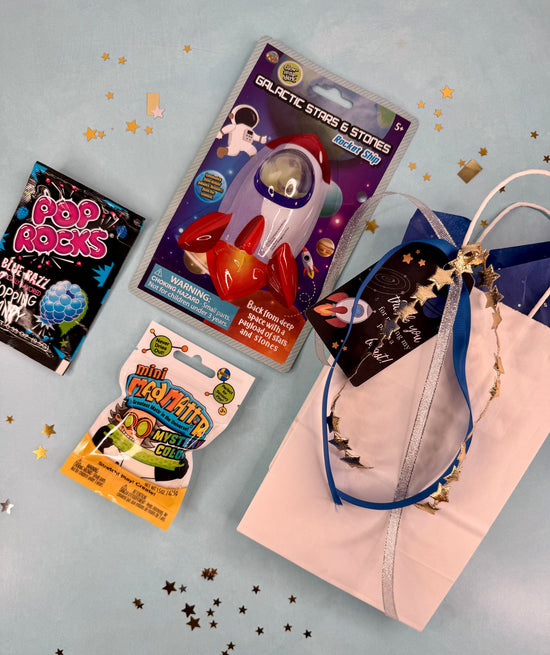 Assorted party favors including a rocket-shaped toy, Pop Rocks, and a lanyard on a gray background with star confetti.