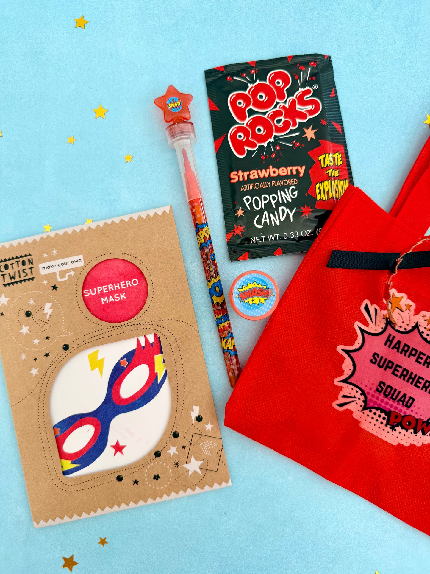 A themed pop-up candy and mask set on a light blue background, featuring a cardboard superhero mask pack, a bag of Pop Rocks strawberry popping candy, a colorful pencil, a round sticker, and a red canvas 'Harper's Superhero Squad' tote bag.