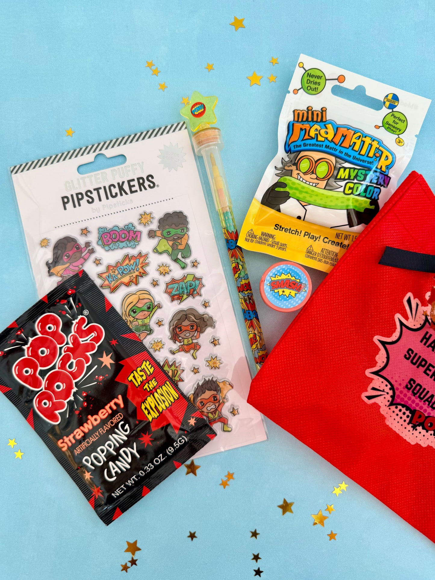 A curated mini superhero gift set arranged on a light blue background with gold star confetti. The set includes a red zip pouch, a pack of colorful superhero stickers, a pack of mini pop rocks candy, a mini plastic toy sword, and a round tin of gum.