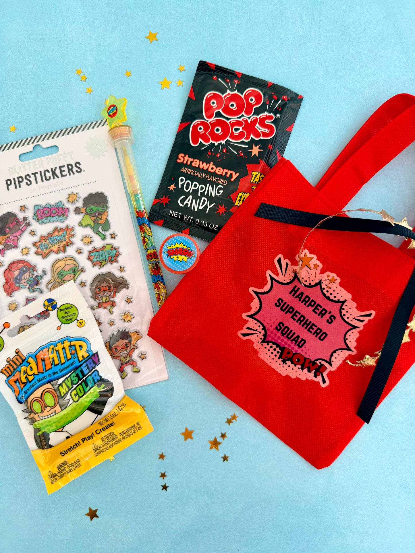 A red canvas tote bag with black handles and a pink comic-style patch reading 'Harper's Superhero Squad 2026' is arranged on a light blue background with gold star confetti. Surrounding the tote are sticker sheets, a Pop Rocks candy packet, a small novelty pen, and a yellow character snack bag.