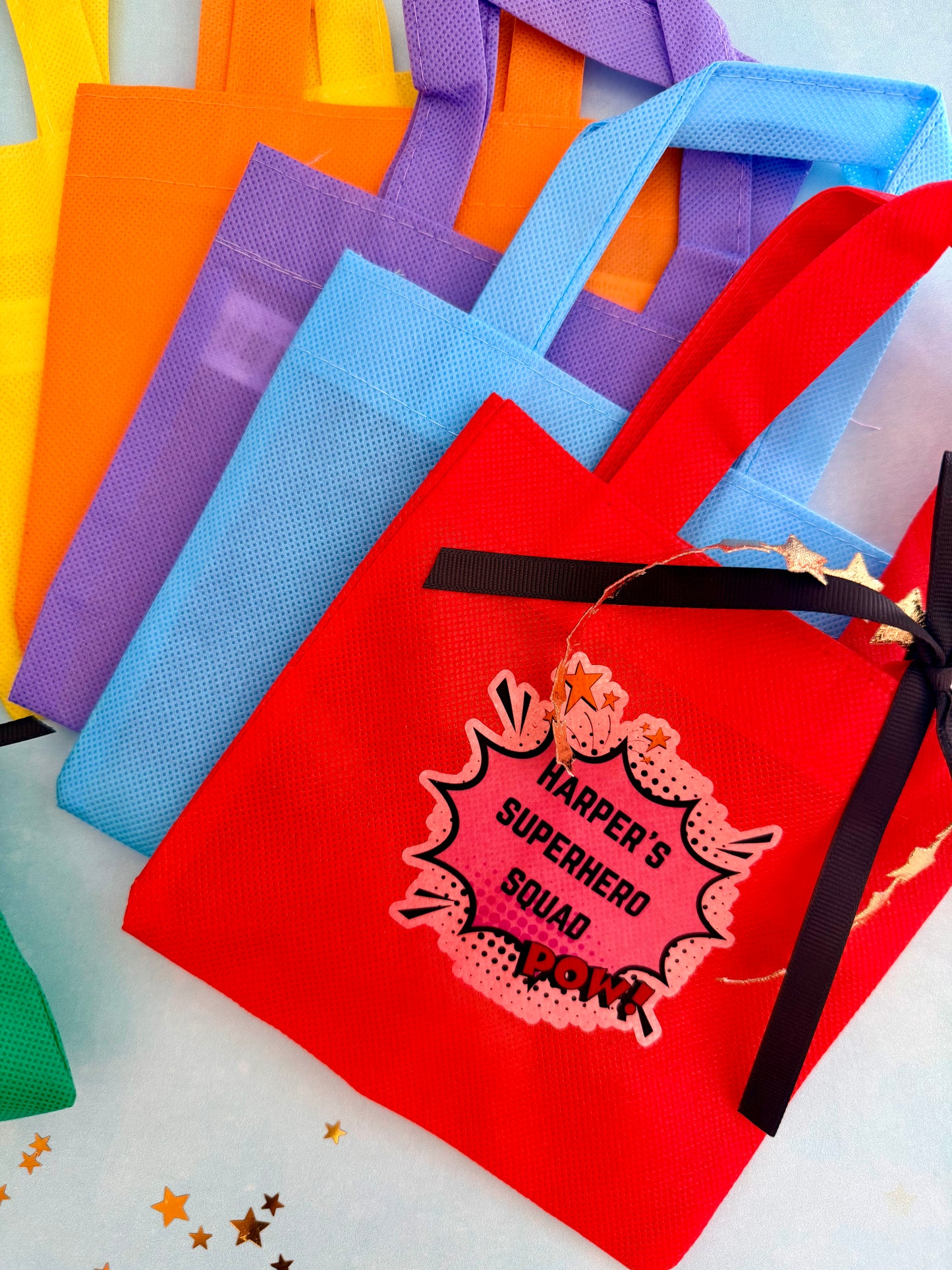 A stack of colorful reusable tote bags in red, blue, purple, orange, and yellow, with the front red bag featuring a black ribbon bow and a comic-style patch that reads 'HARPER'S SUPERHERO SQUAD LOVE'.