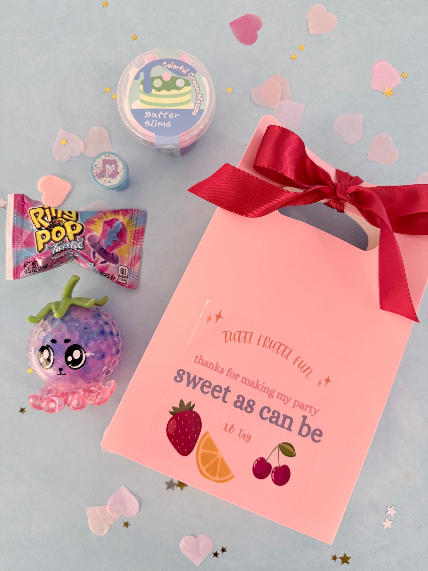 Pink party favor bag with red ribbon, candy, and decorations on a light gray background