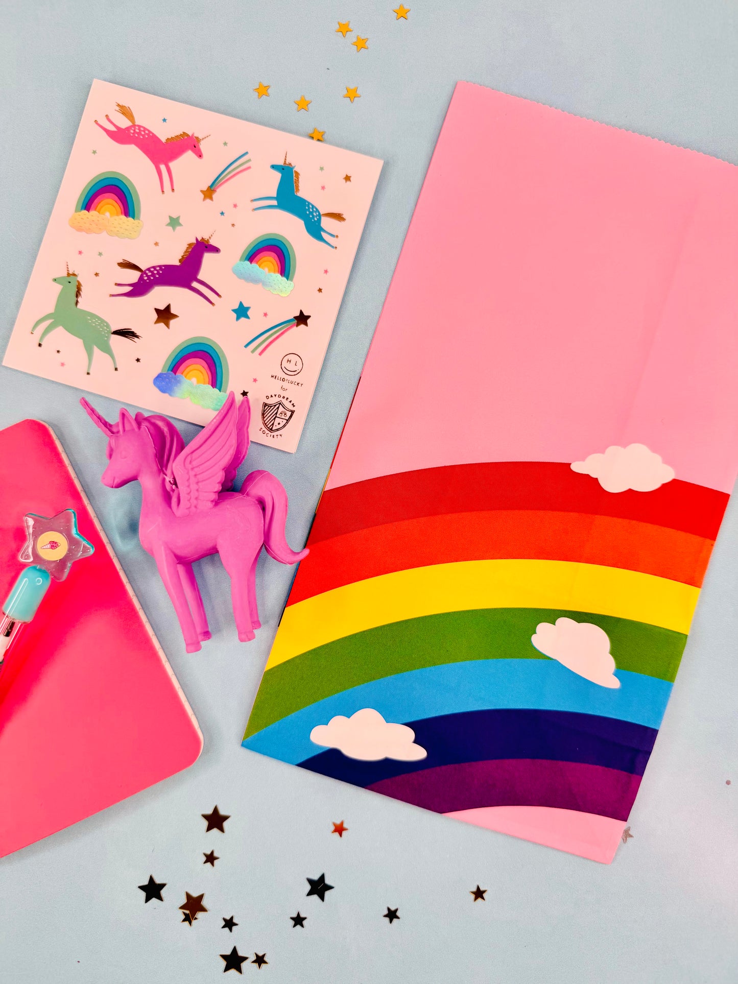 Colorful stationery items including a pink unicorn figurine, rainbow notebook, and star-shaped stickers on a light blue background.