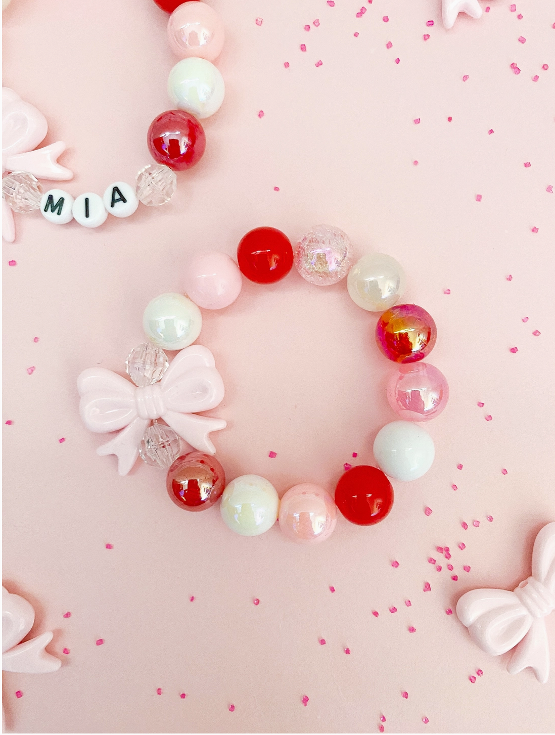 A pink and red acrylic bead bracelet with a bow detail, displayed on a pink background with decorative dots.