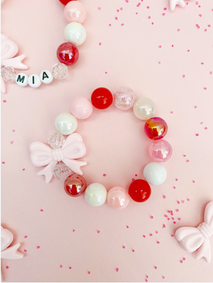 A pink and red acrylic bead bracelet with a bow detail, displayed on a pink background with decorative dots.
