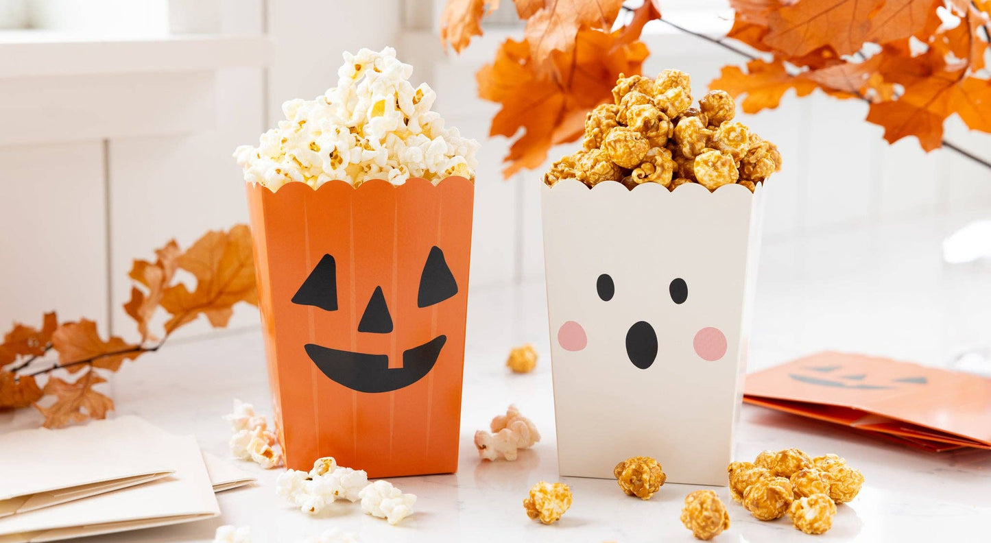 Two Halloween-themed popcorn boxes filled with popcorn on a white tabletop, surrounded by autumn leaves. One box is orange with a jack-o'-lantern face, and the other is white with a ghost face.