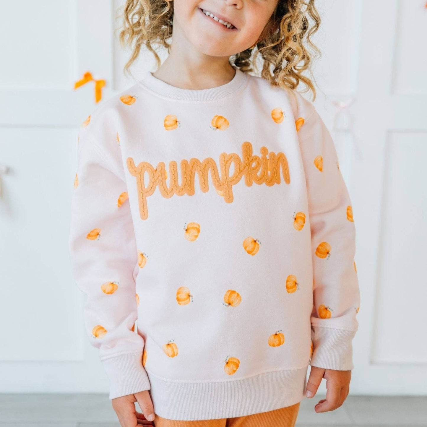 Child wearing a white sweatshirt with orange 'Pumpkin' text and pumpkin patterns on a light background