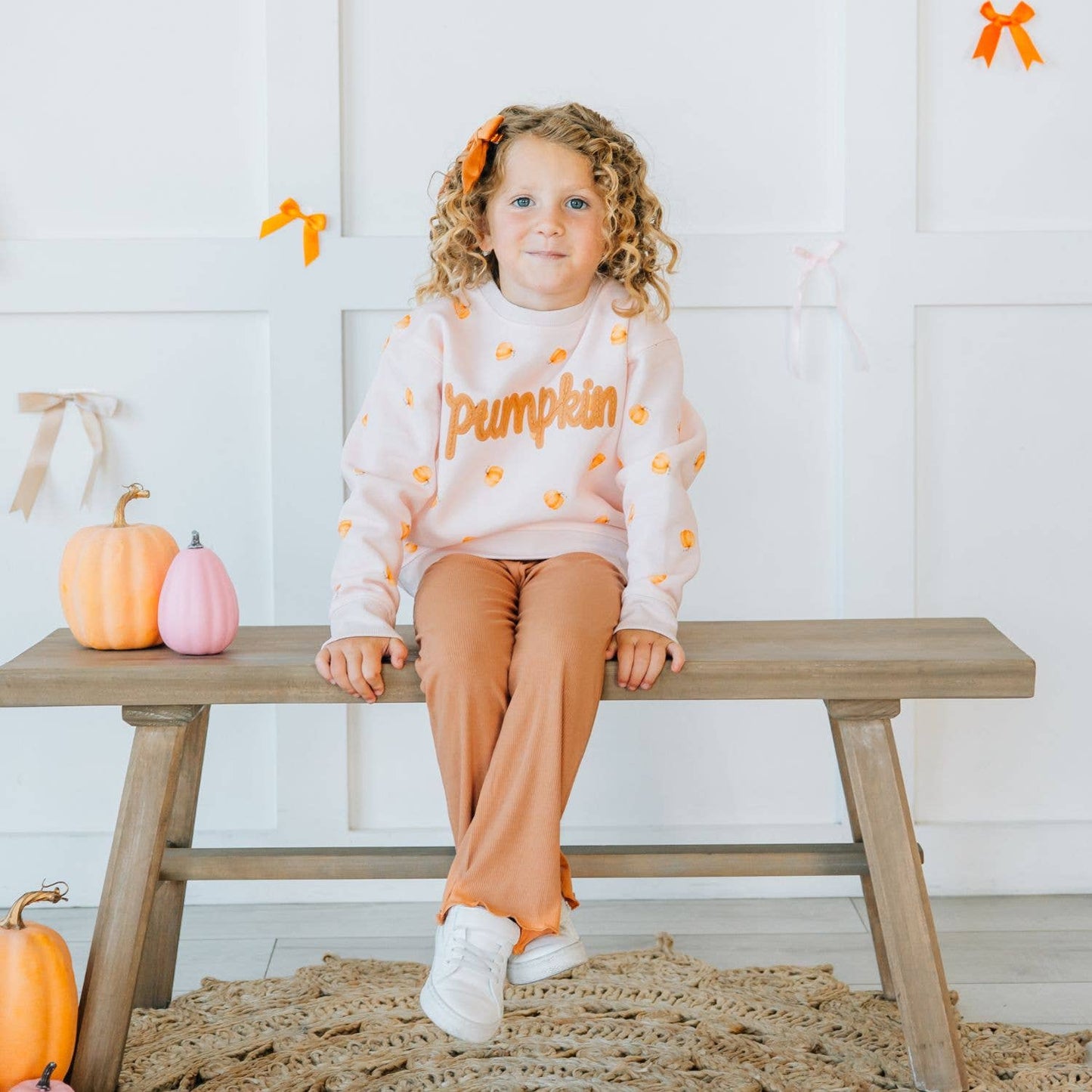 A young girl with curly blonde hair sits on a wooden bench wearing a light pink 'Pumpkin' sweatshirt with orange pumpkin motifs and matching orange-brown pants. The scene is decorated for autumn with small pumpkins, orange ribbons, and a woven rug.