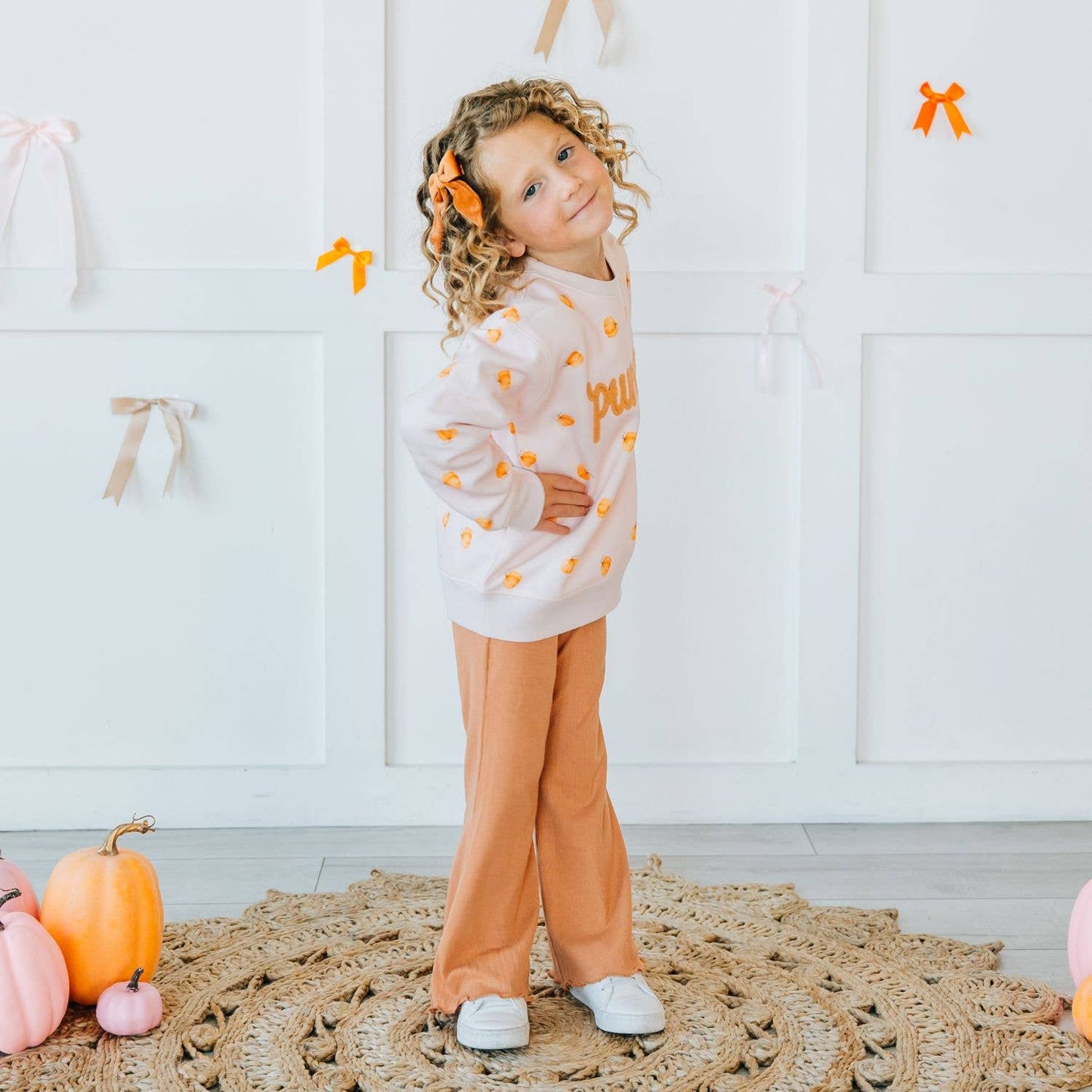 A young girl with curly hair stands on a woven rug in a white room decorated with pumpkins and ribbon bows. She is wearing a white long-sleeve sweatshirt with orange pumpkin prints and orange lettering, paired with orange pants.
