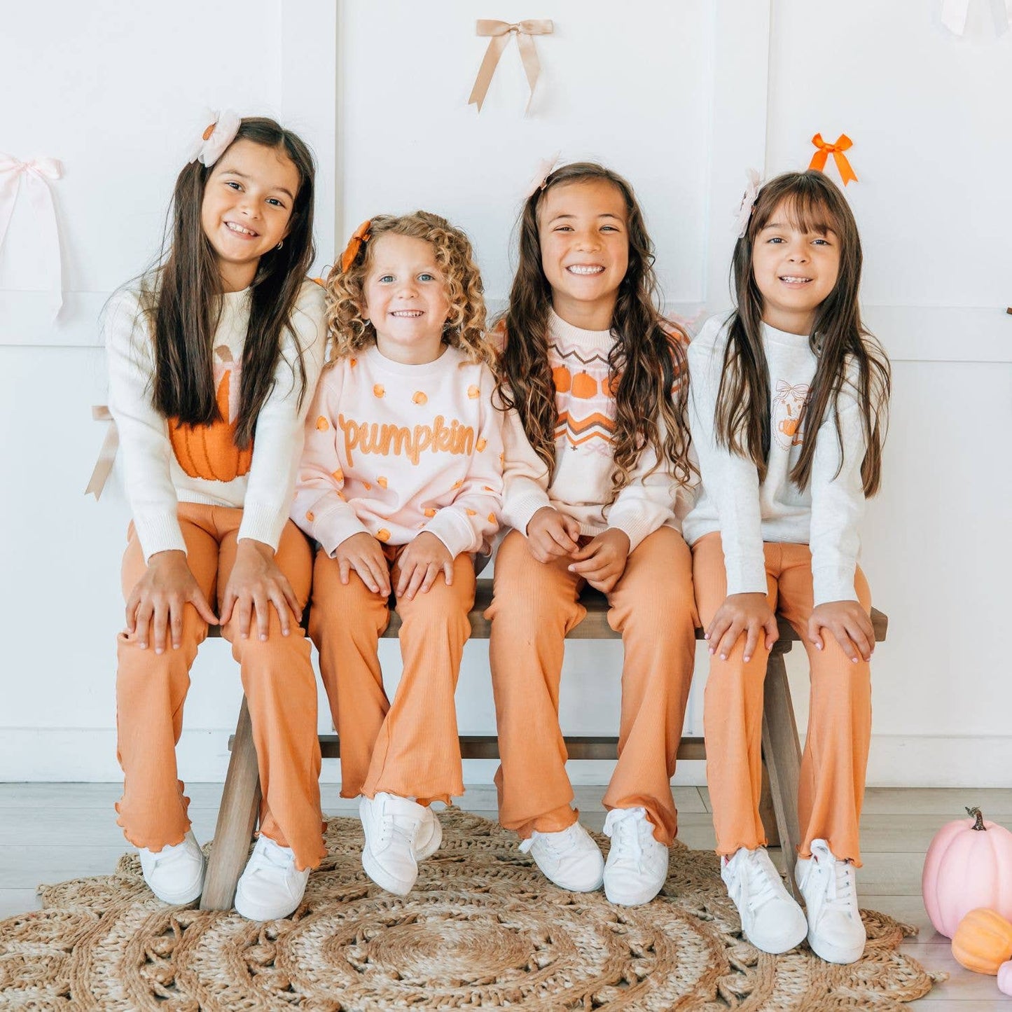 Four young girls sitting together on a bench in a bright, white room, wearing matching autumn-themed sweatshirts and sweatpants in orange, peach, and white.