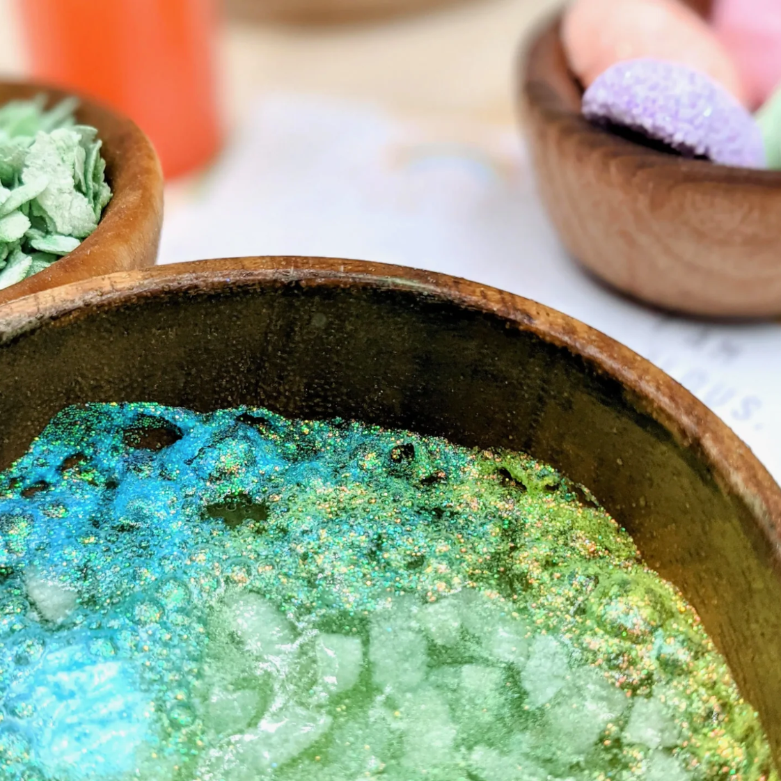 A close-up of a wooden bowl filled with sparkling blue and green glittery powder or bath salts, with other bowls blurred in the background.