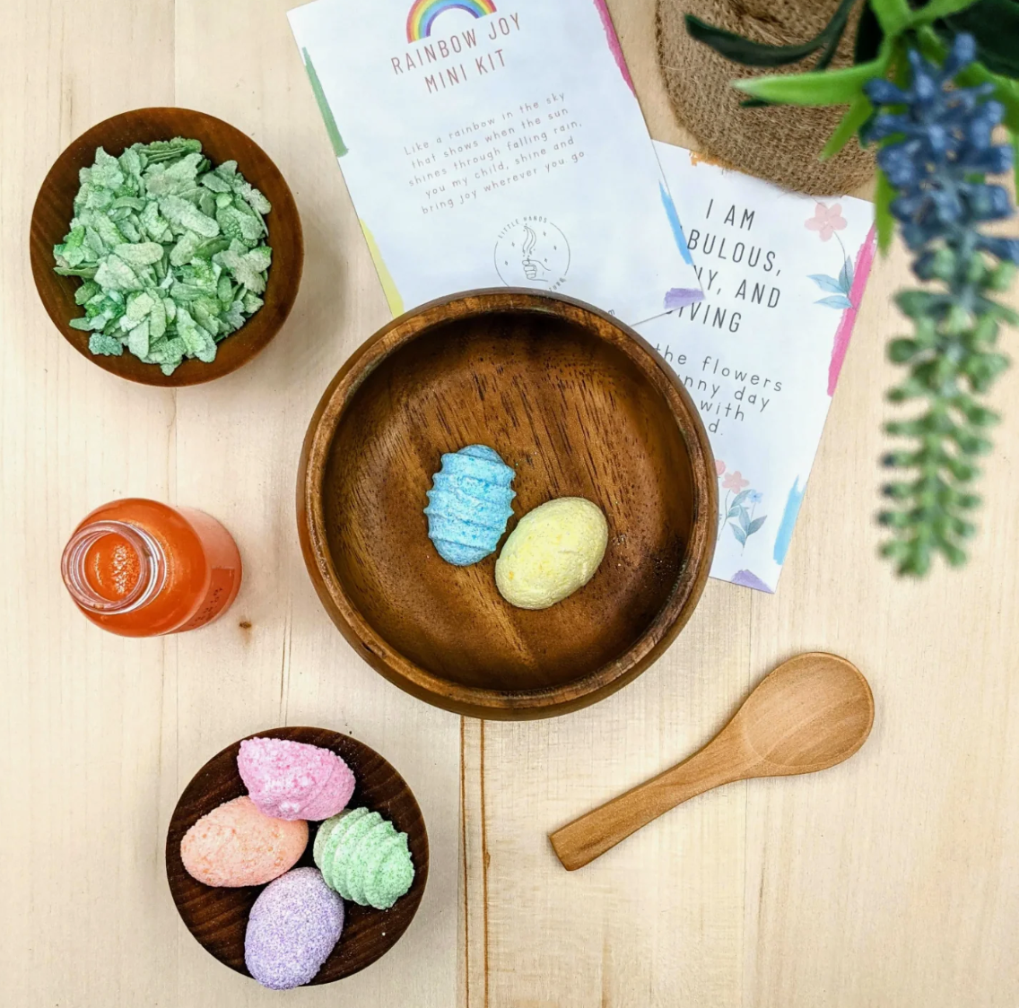 A styled flat-lay of a 'Rainbow Joy Mini Kit' on a light wood background, featuring colorful bath bombs, bath salts, a small glass of orange liquid, a wooden spoon, and printed cards.