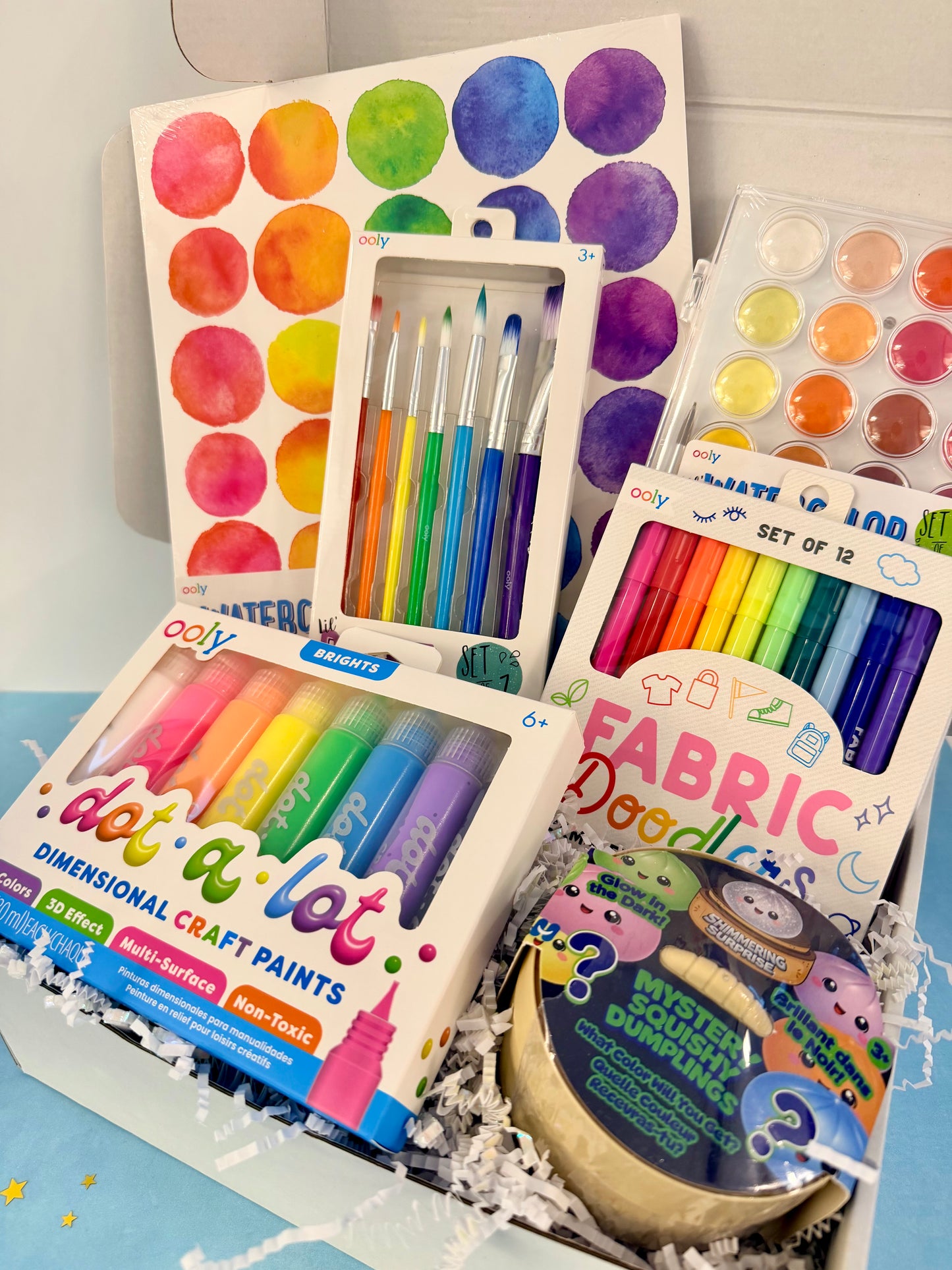 A curated craft and art gift box filled with colorful items, including a watercolor paint palette, fabric markers, dimension craft paints, paintbrushes, and a mystery gem package, arranged in white paper shred inside a box.