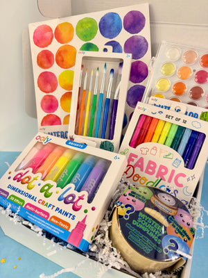 A curated craft and art gift box filled with colorful items, including a watercolor paint palette, fabric markers, dimension craft paints, paintbrushes, and a mystery gem package, arranged in white paper shred inside a box.