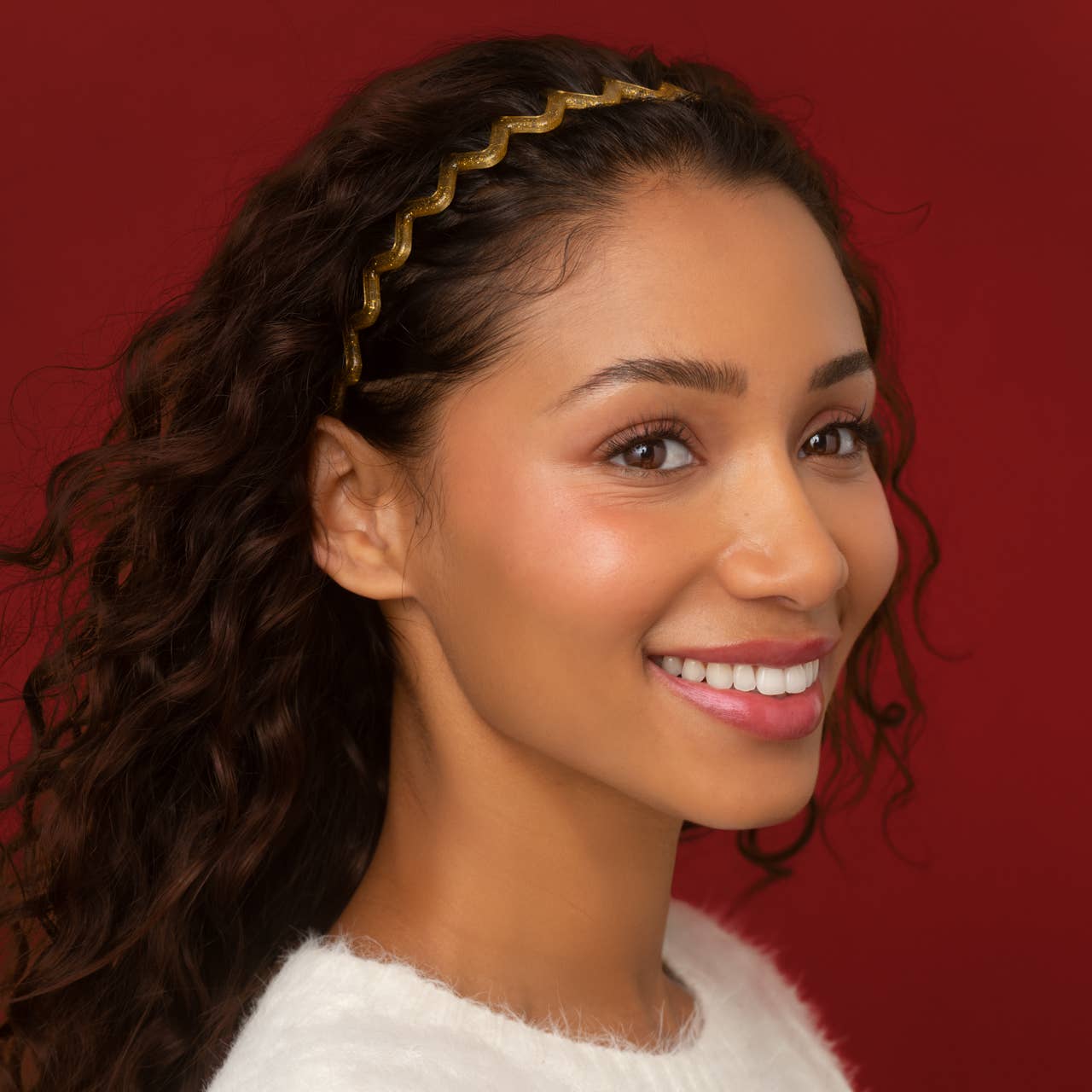 A smiling woman with long dark curly hair wears a gold wavy headband against a solid red background.
