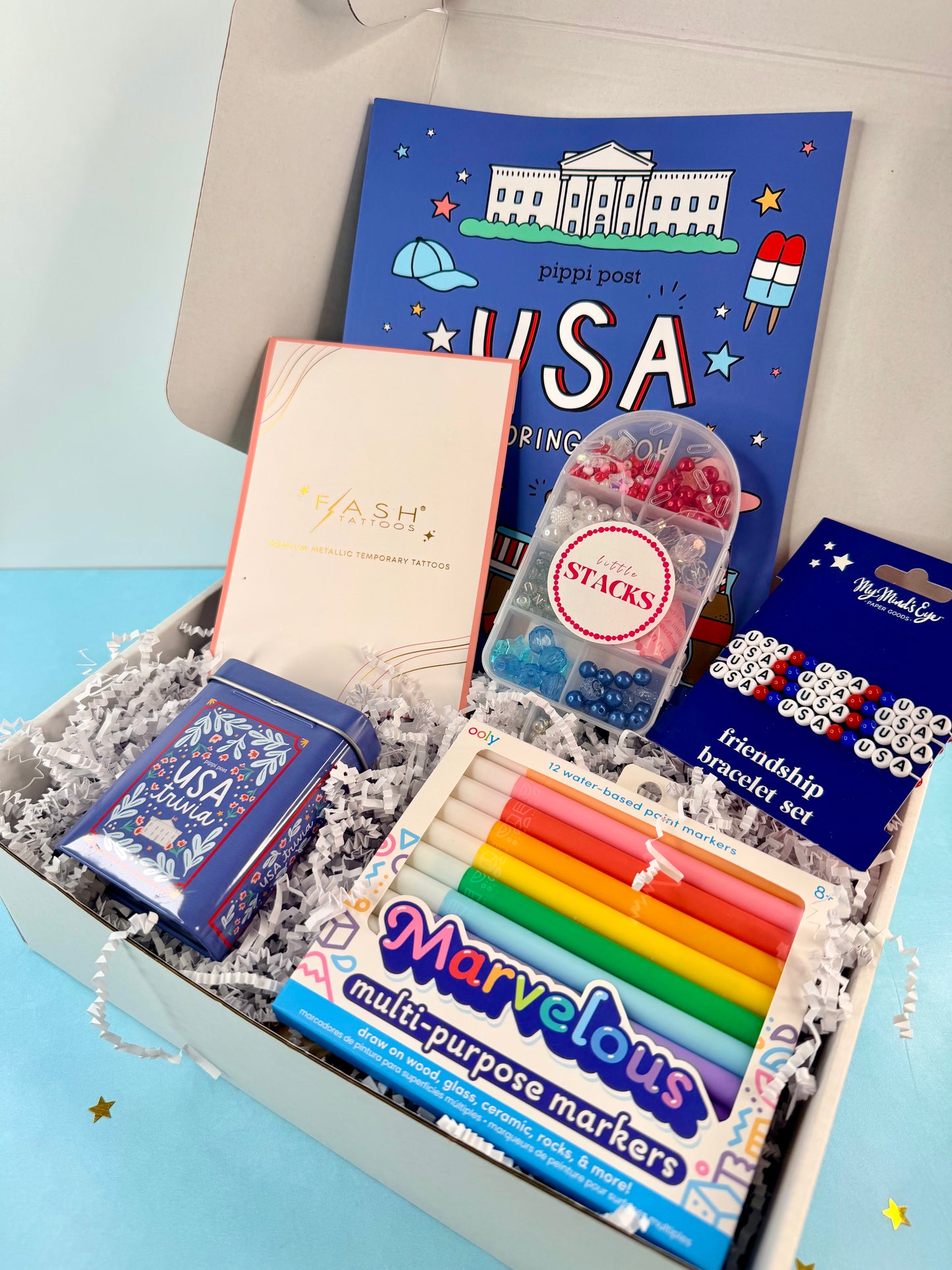 Gift box with stationery items including markers, a book, and small containers on a light blue background.