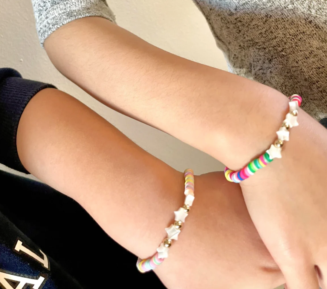 Two beaded bracelets with white star charms worn on a person's wrists. One bracelet features pastel pink, blue, and yellow beads, while the other has multicolor beads including pink, green, blue, and white.