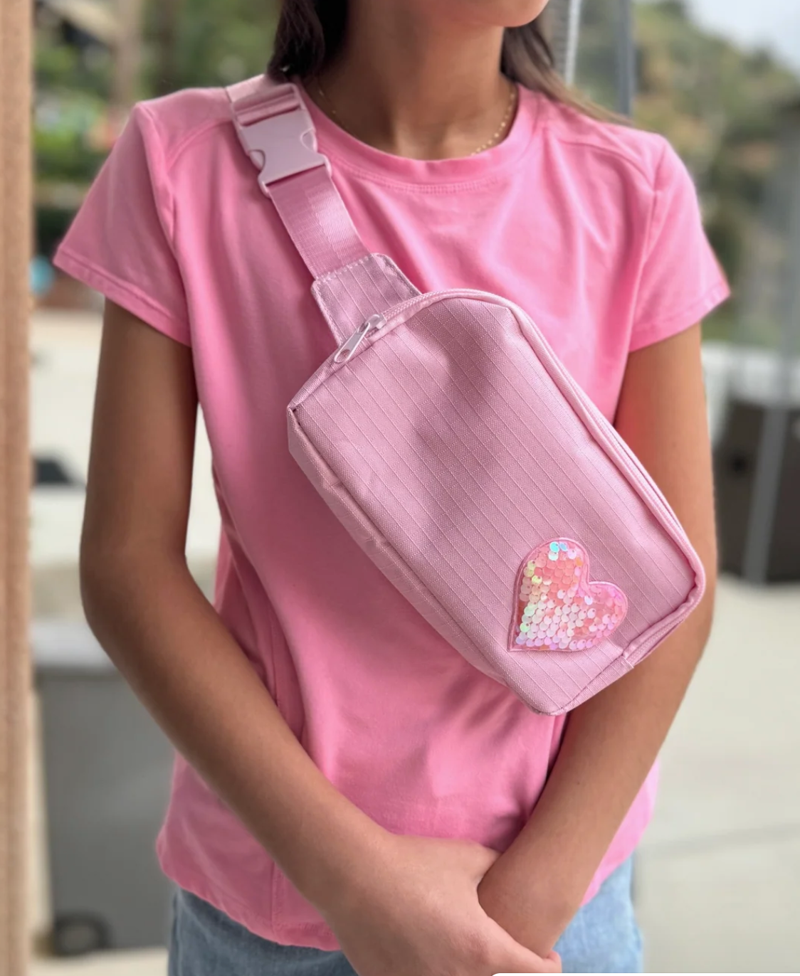 A pink nylon sling bag with an adjustable strap and a sequined heart patch, worn by a person in a pink t-shirt.