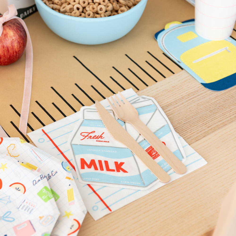 A styled tabletop scene with a milk carton printed napkin as the main product, accompanied by wooden utensils. The napkin has a retro milk carton design with red and blue lettering on a white background.