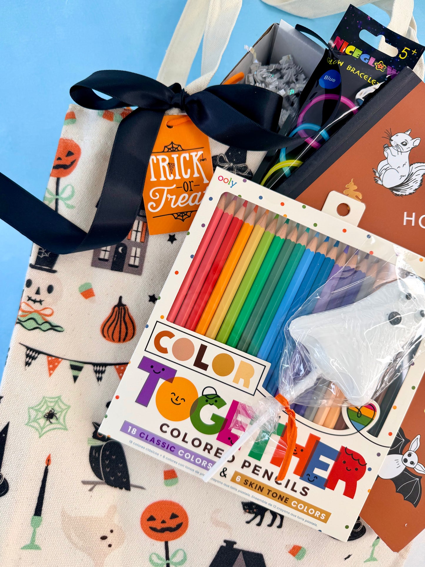 A Halloween-themed gift basket or bag filled with stationery and treats, featuring a printed canvas tote with a black ribbon, a box of rainbow-colored pencils labeled 'Color Together', a ghost-shaped eraser, and a 'Trick or Treat' tag against a light blue background.