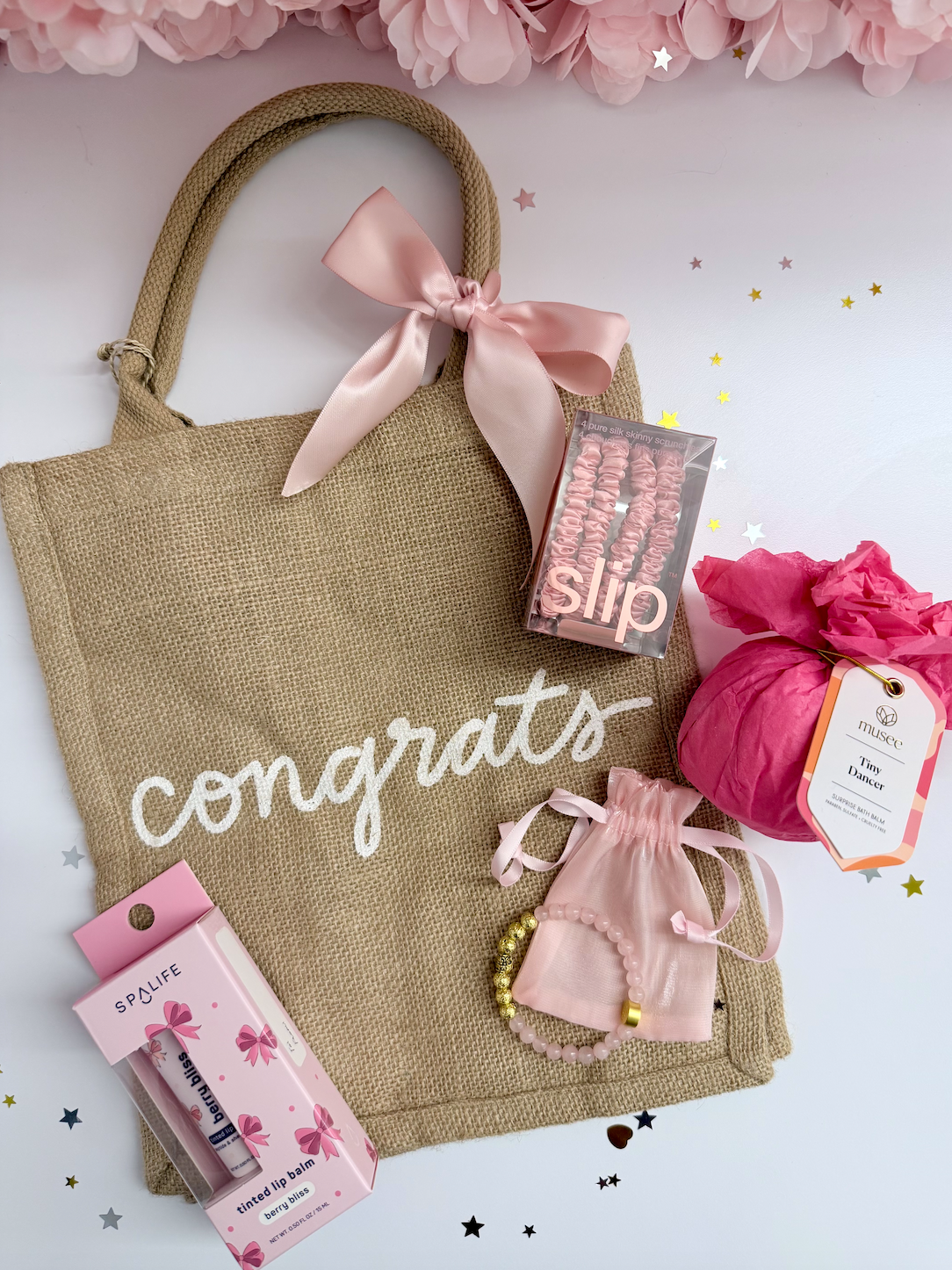 Gift bag with 'congrats' text, pink ribbons, and small items on a white background with pink flowers.