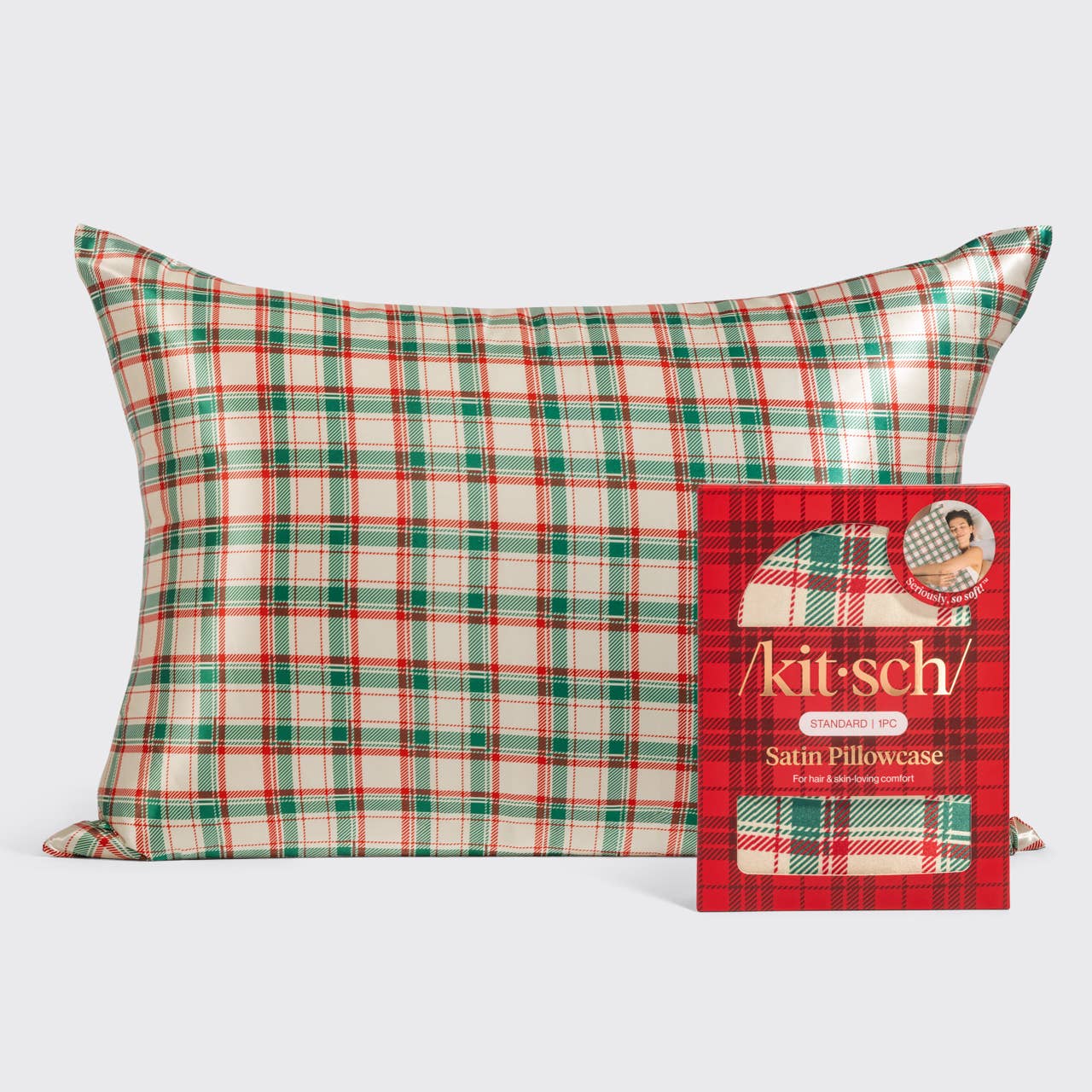 A rectangular satin pillowcase with a red, green, and cream plaid pattern, shown next to its red branded retail box on a light gray background.