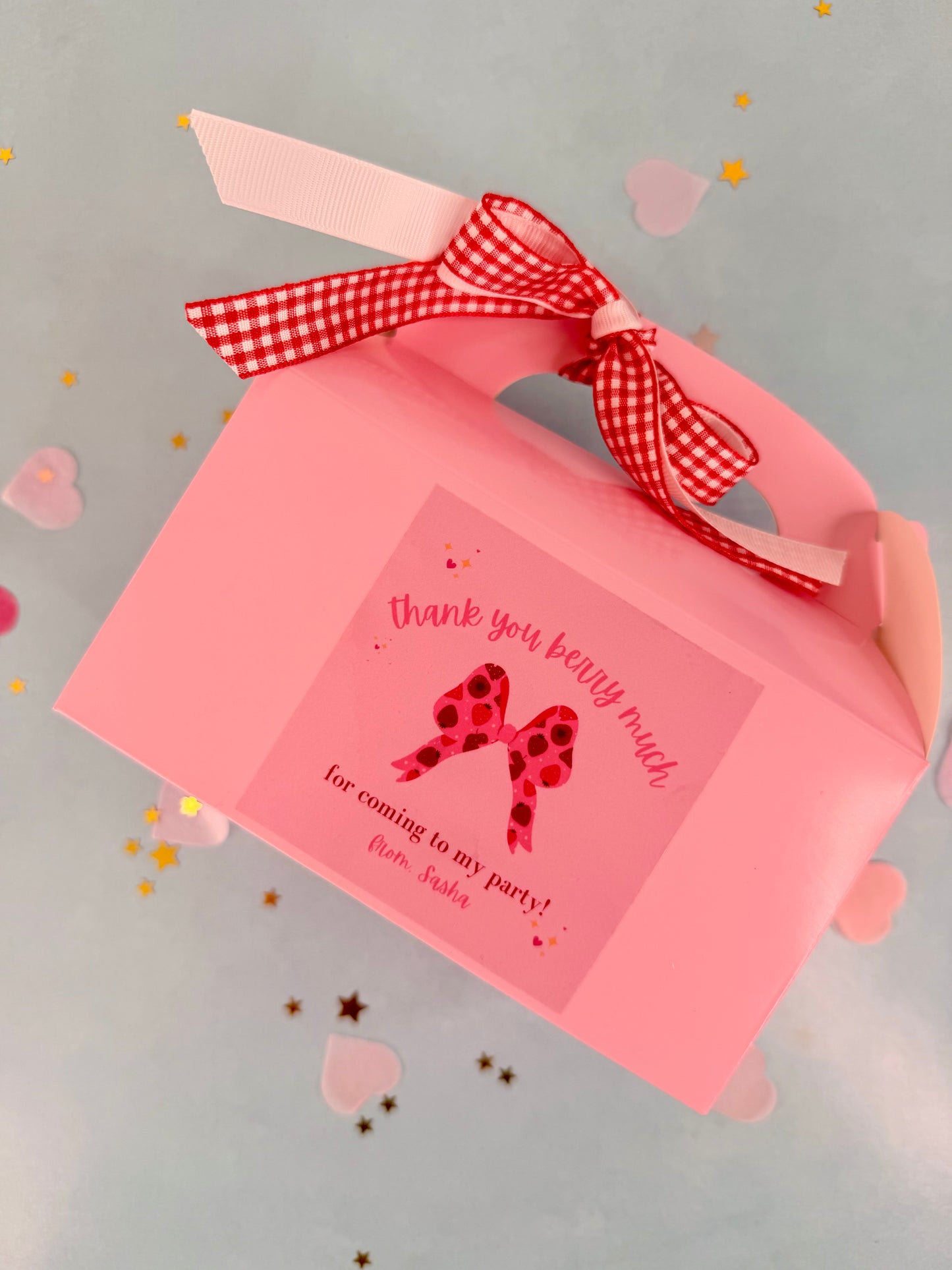 Pink party favor box with a red checkered ribbon and a card inside on a gray background.