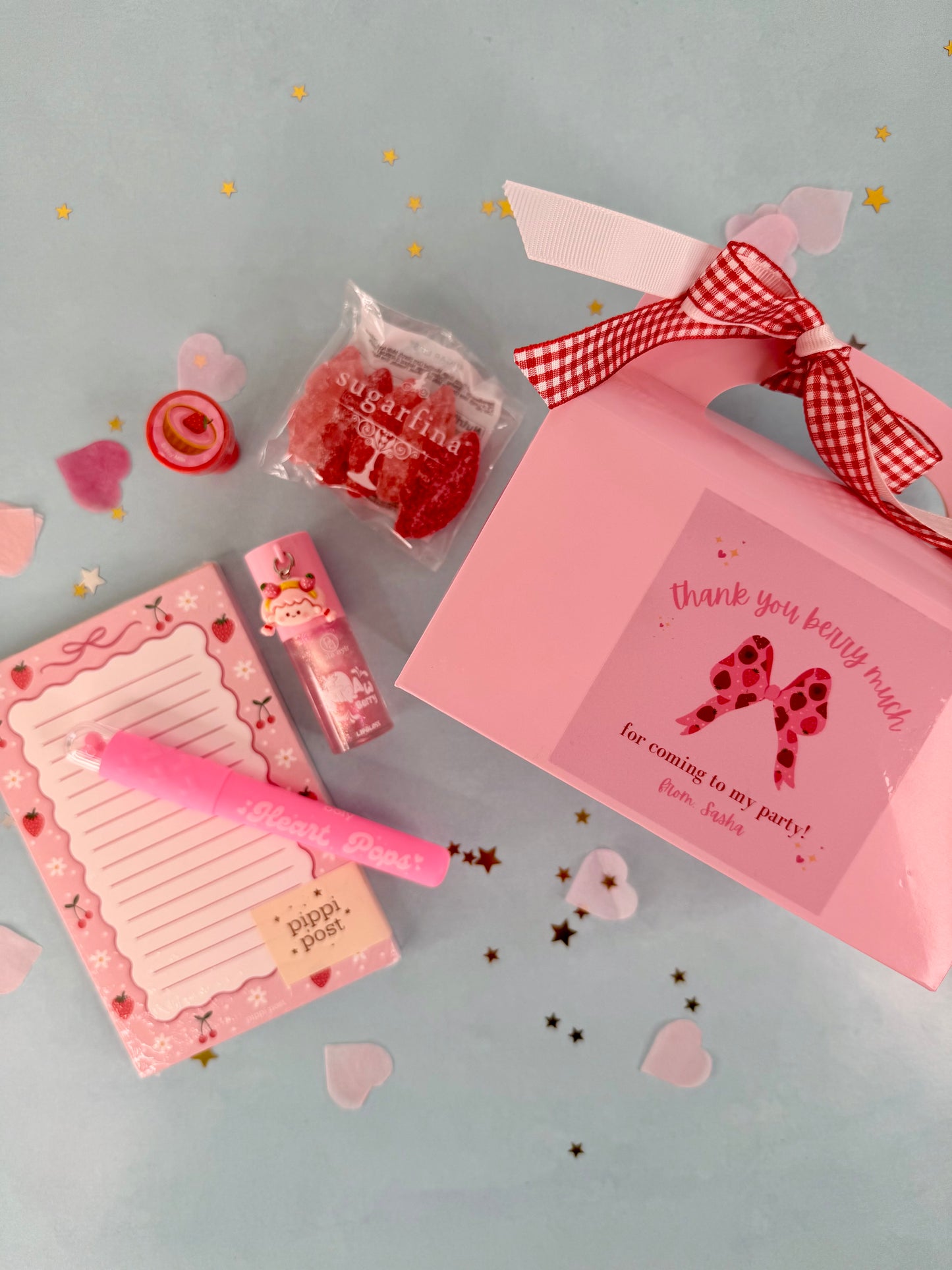 Pink party favor set with a card, candy, and pen on a light blue background.