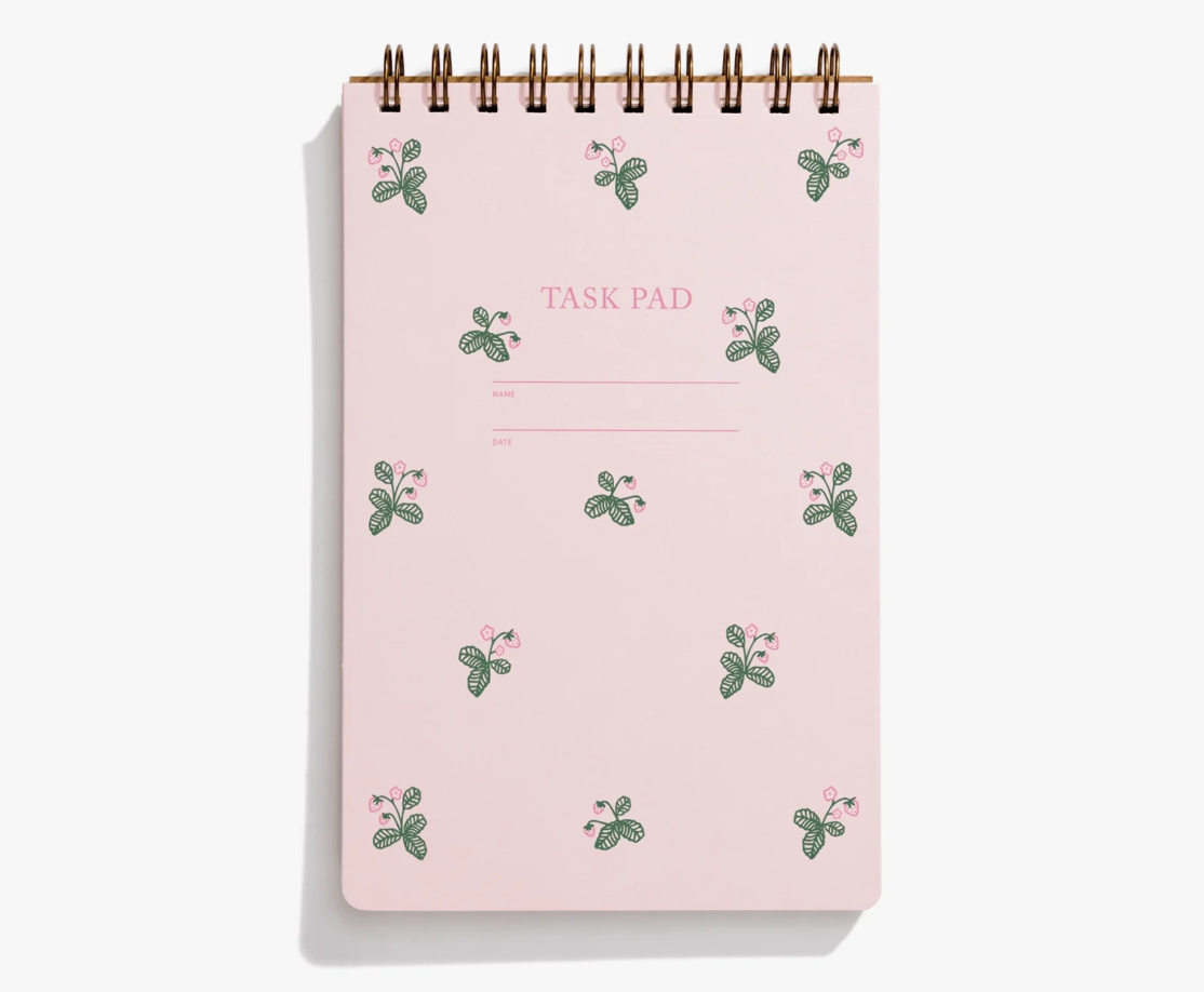 A spiral-bound task pad with a light pink cover decorated with small green leaves and pink flowers, featuring the text 'TASK PAD' in the center.