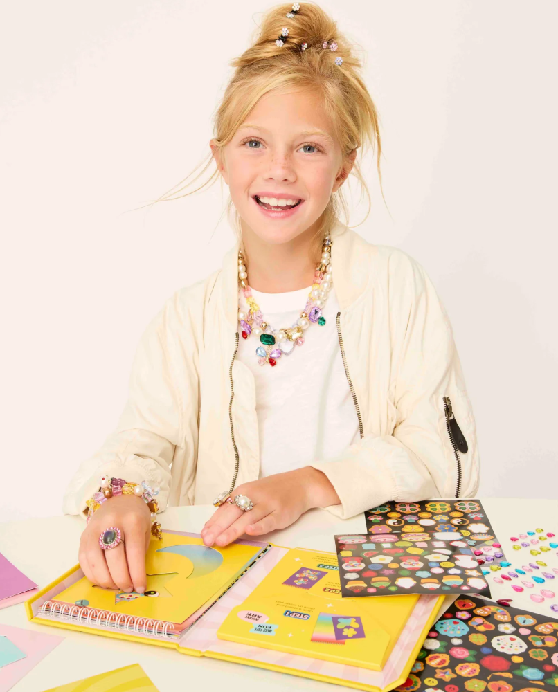 Young girl with a colorful necklace and bracelets sitting at a table with craft materials.