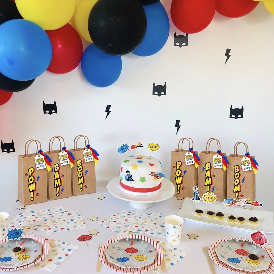 A themed party setup with Batman decorations, including a white and red decorated cake on a stand, brown paper gift bags with 'POW! BAM! BOOM!' tags, matching party plates, cups, and napkins, and a wall decorated with black bat silhouettes and colorful balloons.