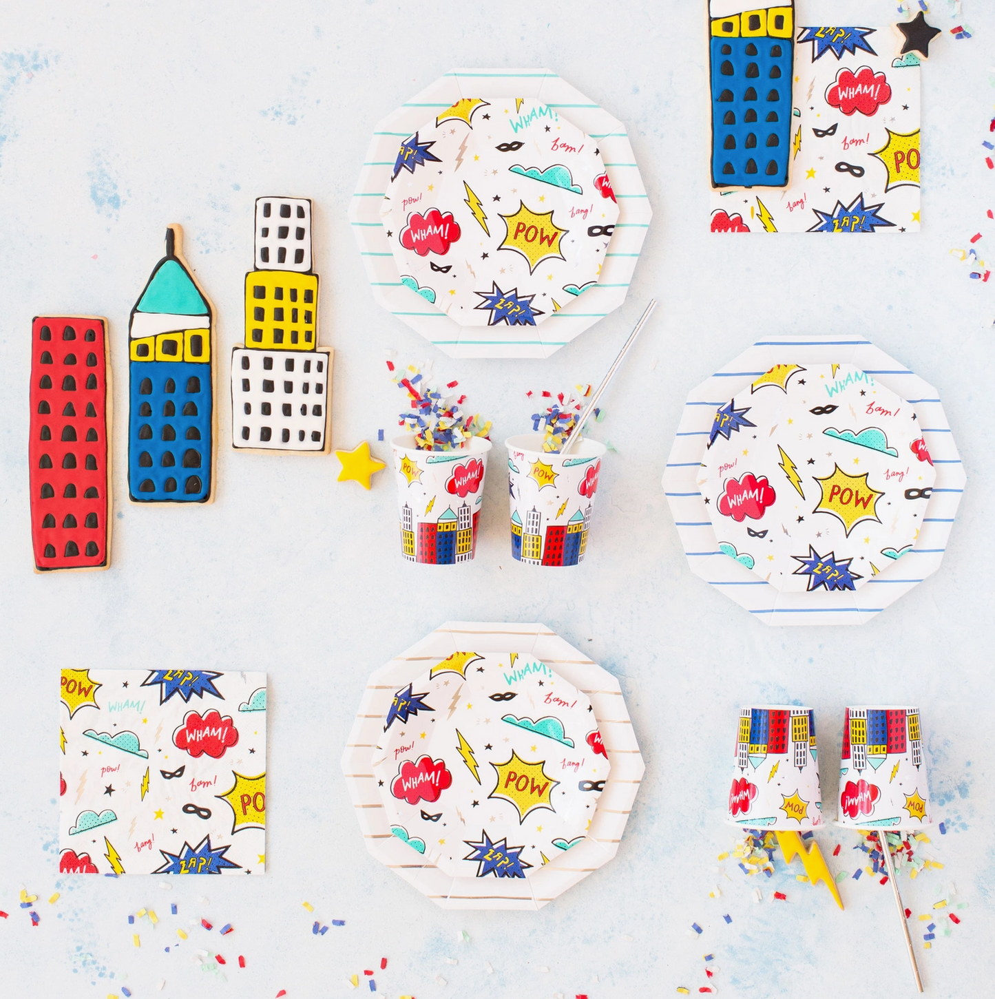 Comic book themed party tableware set including paper plates, cups, napkins, and cookie cutters with colorful pop-art illustrations, stars, and speech bubbles on a light blue background.