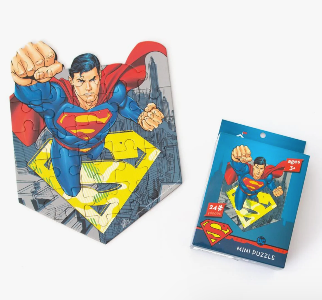 A Superman-themed mini jigsaw puzzle shown partially assembled beside its blue retail box on a clean white background. The puzzle features Superman flying above a large yellow 'S' emblem with a gray cityscape behind him.