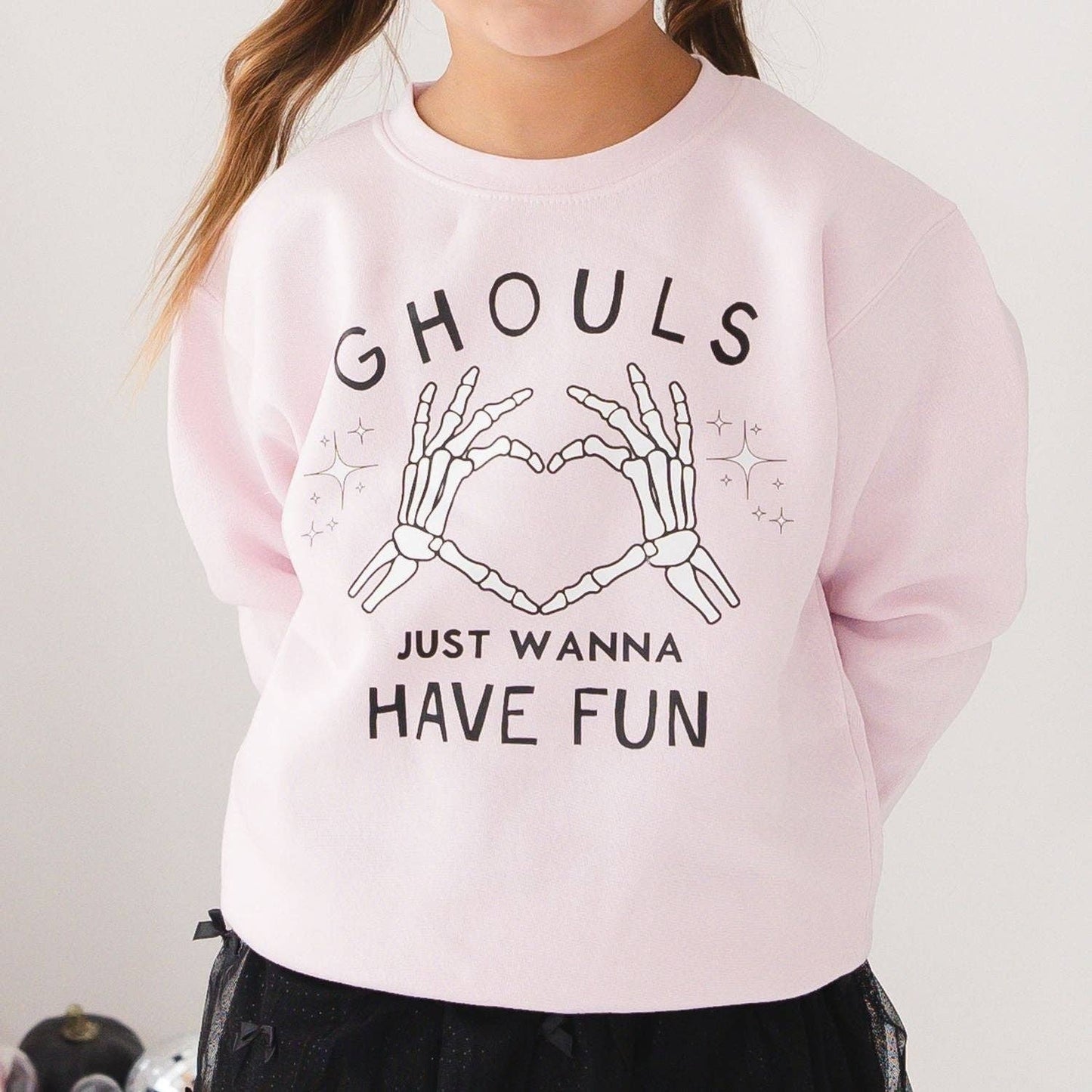 A child wearing a light pink crewneck sweatshirt with a black graphic print that reads 'GHOULS JUST WANNA HAVE FUN' with skeleton hands forming a heart shape.