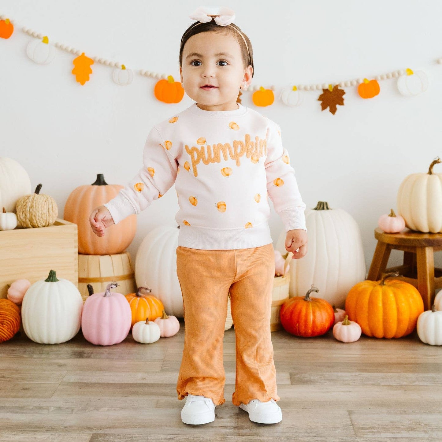 A toddler stands in a decorated fall-themed room wearing a white long-sleeve sweatshirt with orange 'pumpkin' lettering and small pumpkin prints, paired with orange pants and white sneakers.