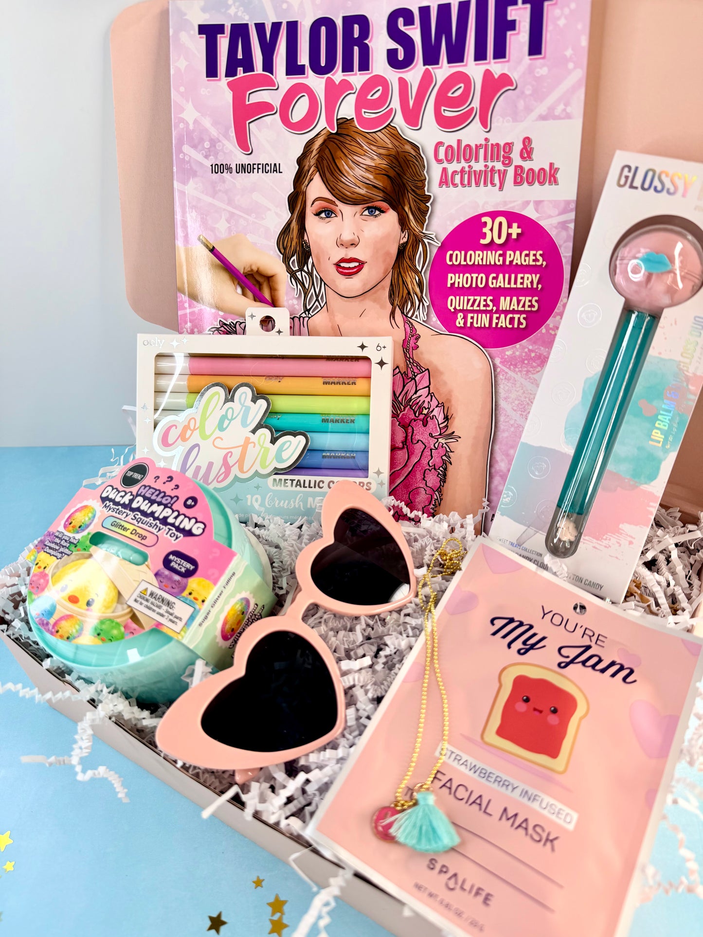 A curated gift box set featuring a Taylor Swift Forever coloring book, a box of glossy lip balm, pink heart-shaped sunglasses, a colorful metallic tin, and a pink facial mask, arranged in white shredded paper.