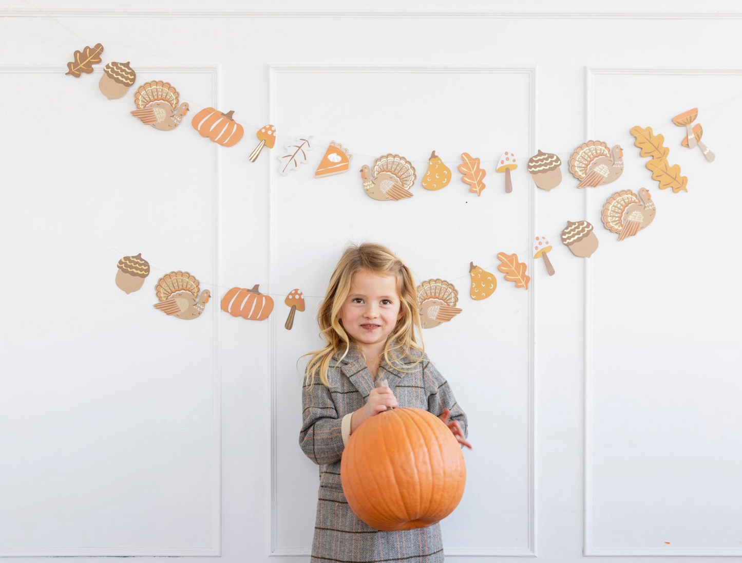 Young girl holding a pumpkin in front of a white wall with autumn-themed decorations.