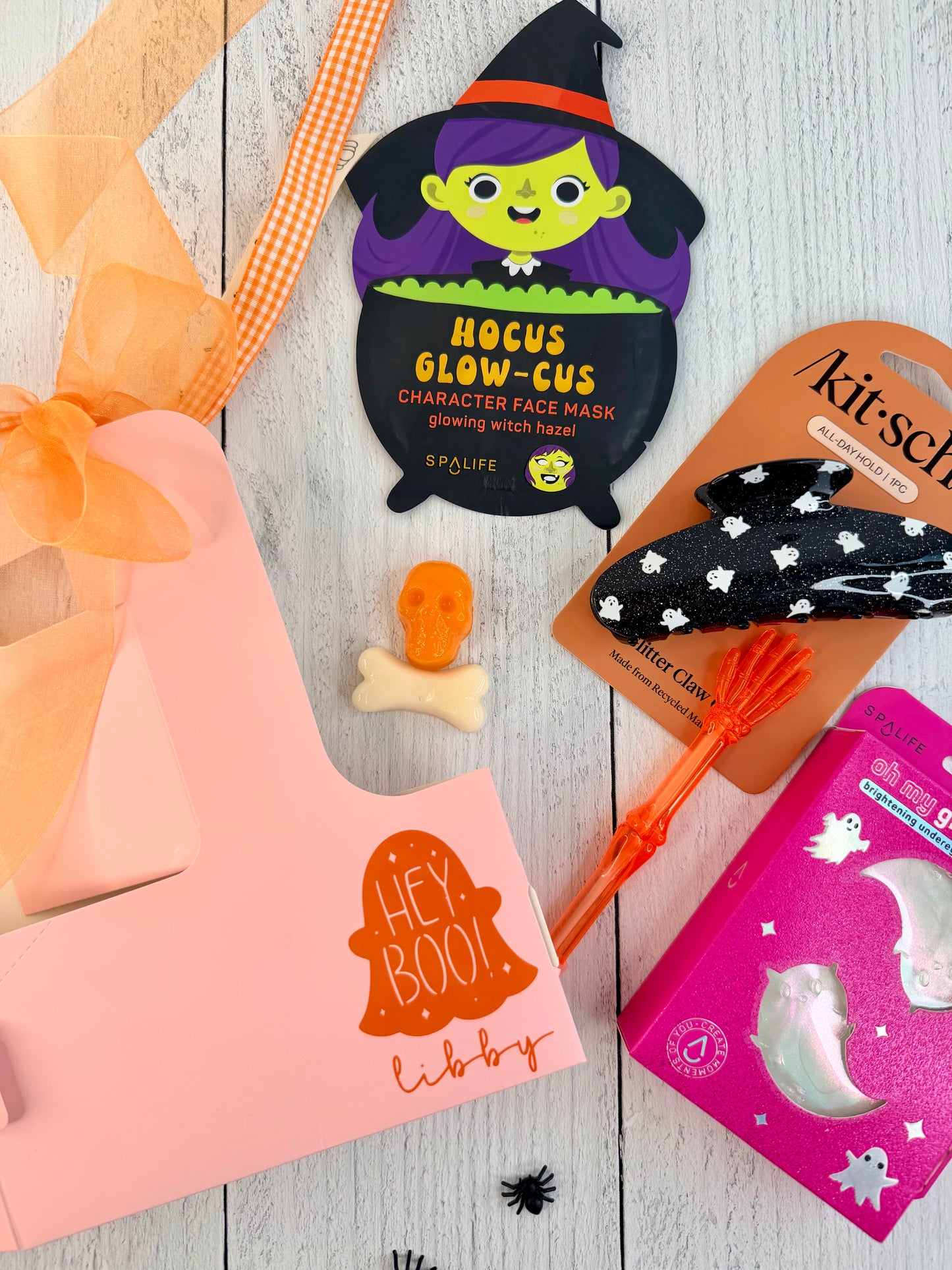 A styled flat-lay of Halloween-themed beauty products on a white wood background, including a pink gift box with orange ribbon, a witch face mask packet, a black star-patterned headband on a card, an orange hairbrush, and a pink ghost box.