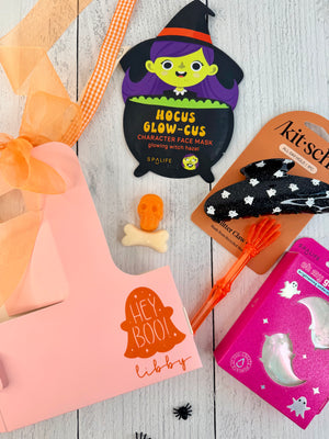 A styled flat-lay of Halloween-themed beauty products on a white wood background, including a pink gift box with orange ribbon, a witch face mask packet, a black star-patterned headband on a card, an orange hairbrush, and a pink ghost box.