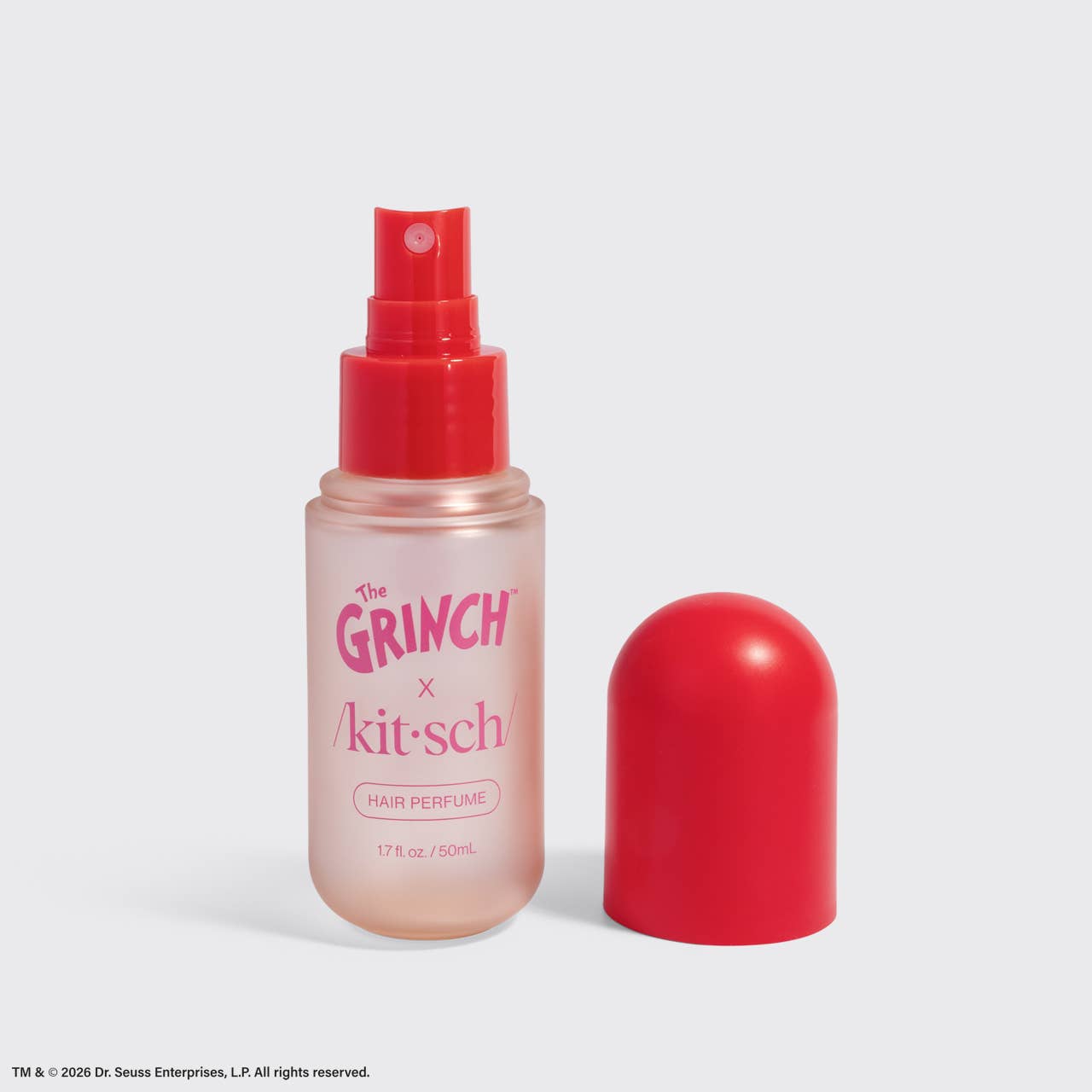 A bottle of Dr. Seuss Enterprises x Kit-Sch Hair Perfume with a translucent pink body and a bright red spray top, shown next to its matching red cap on a light gray background.