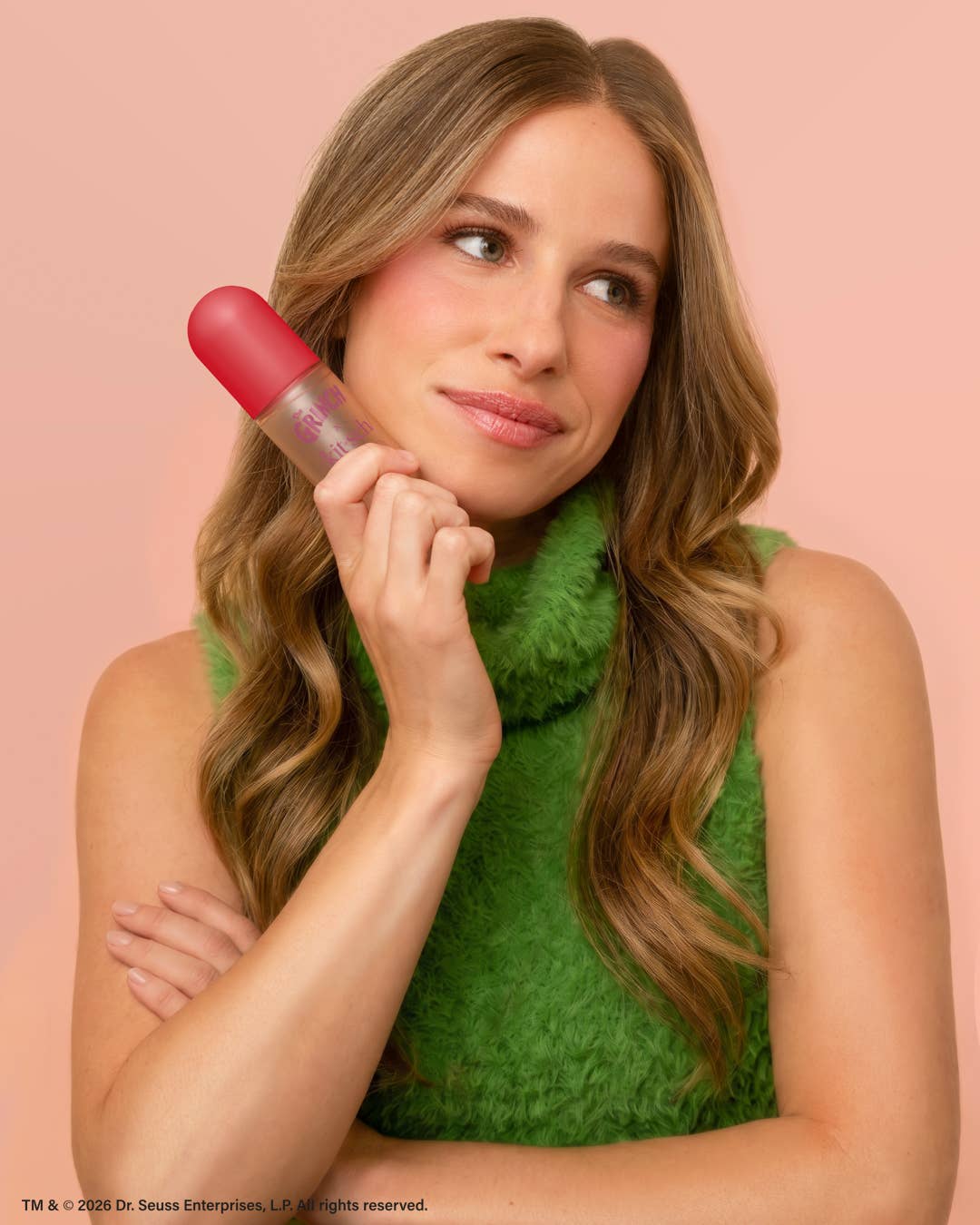 A woman with long wavy light brown hair holds a pink and clear cosmetic tube near her face against a solid pink background. She is wearing a textured green top.