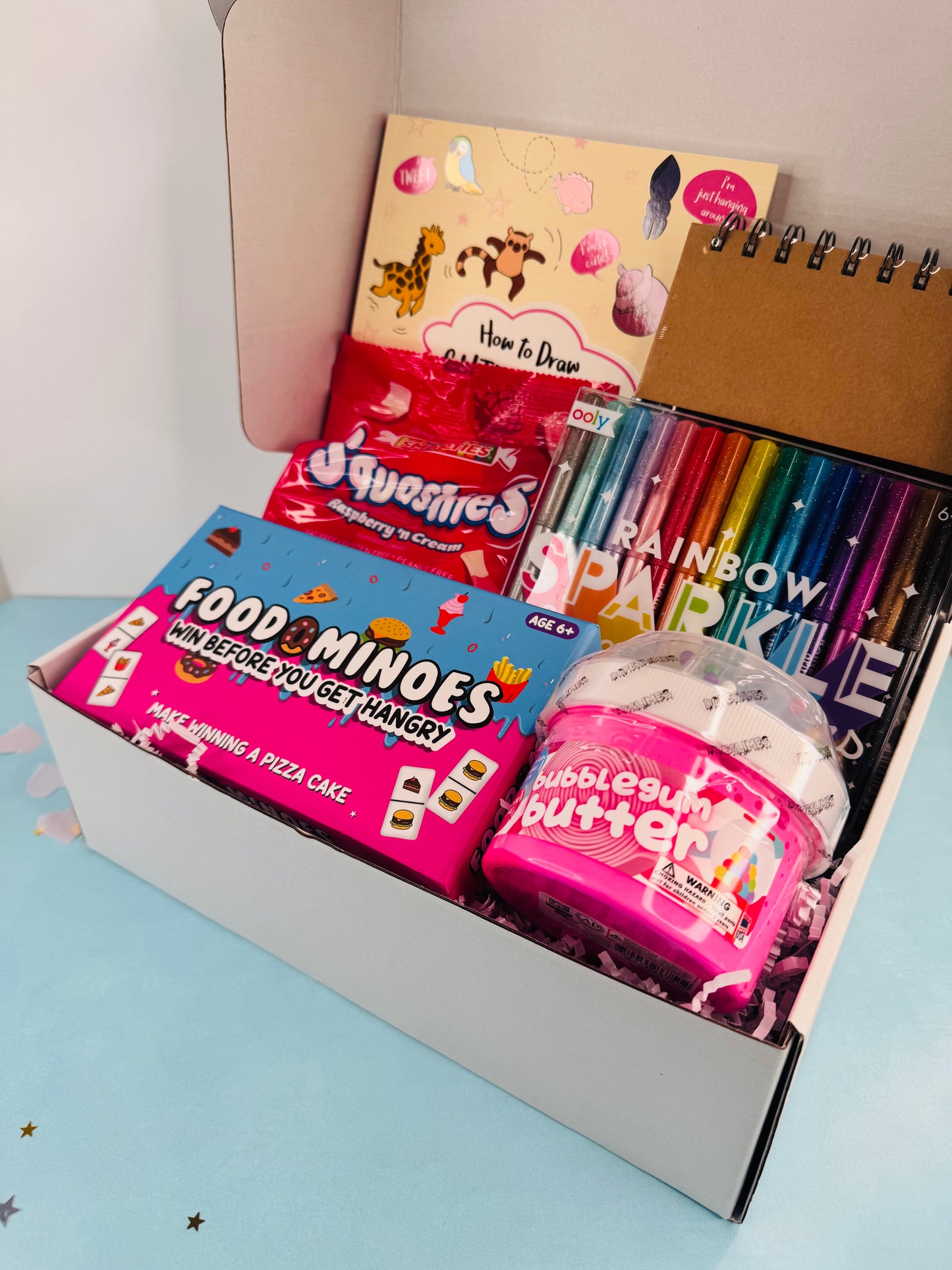 Open box containing various items including a notebook, candy, and a toy, on a light blue surface.
