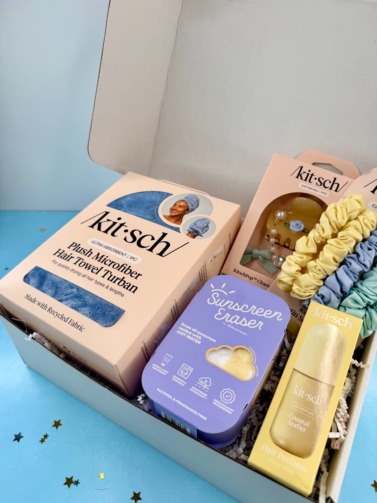Kitsch beauty products including a towel, sunscreen eraser, and serum in a box on a blue background.