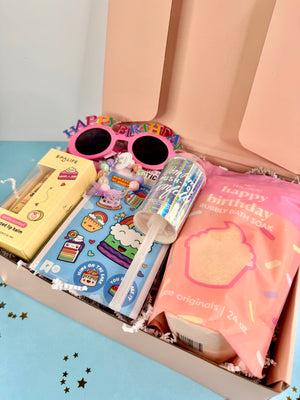 Open box containing birthday-themed items including sunglasses, a book, and soap on a light blue surface.