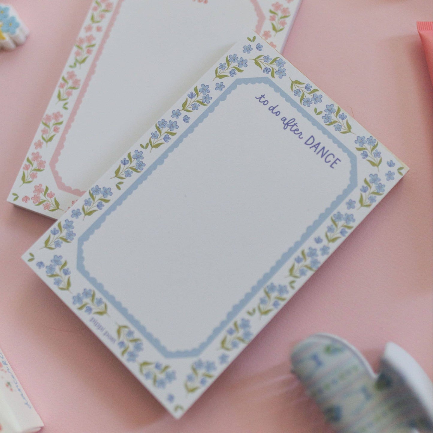 A floral notepad with a blue border and the phrase 'to do after DANCE' in script, placed on a pink surface with other pastel notepads partially visible.