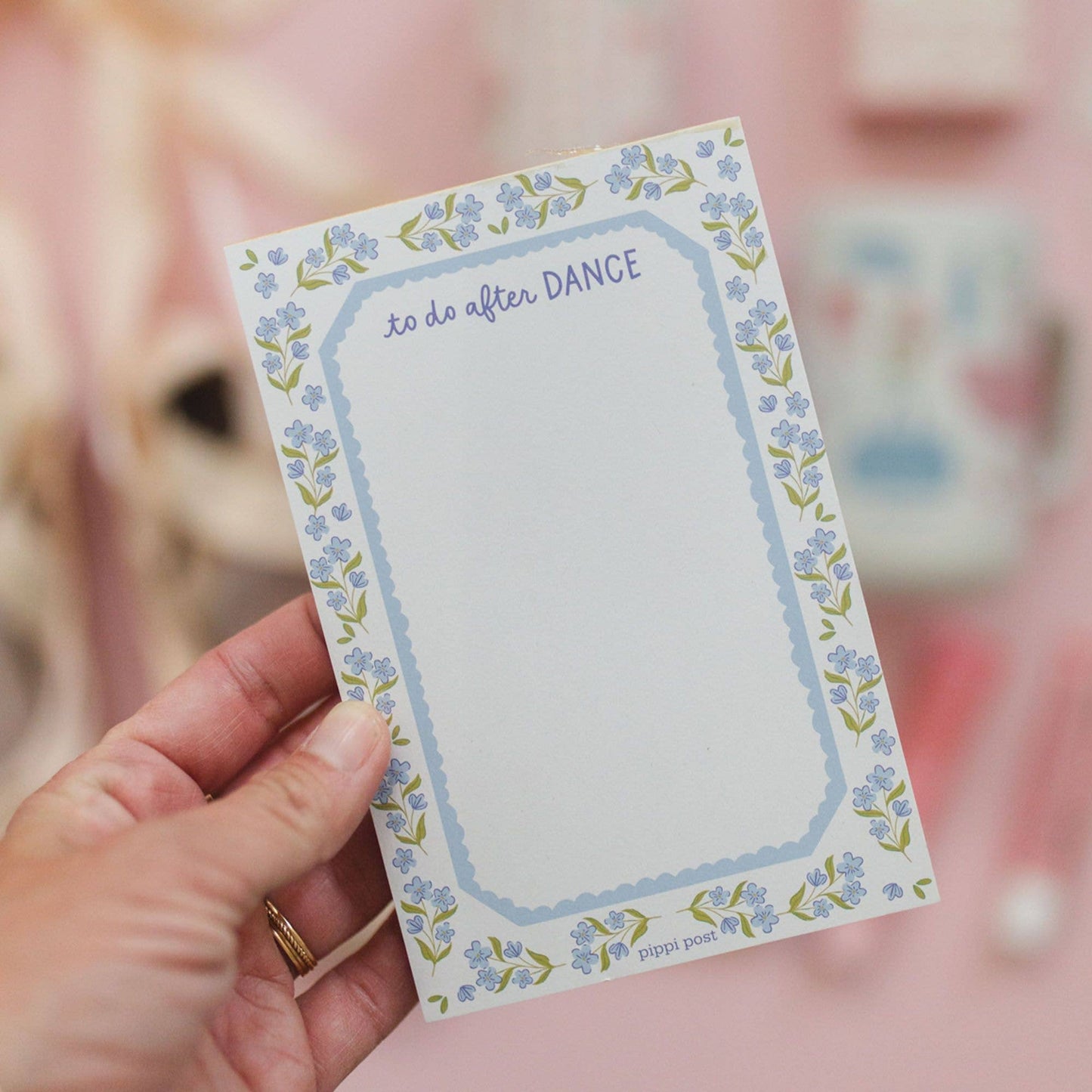 A hand holding a small notepad with a floral border and the phrase 'to do after DANCE' at the top, against a soft pink blurred background.