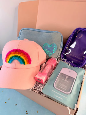 A curated gift box containing a pink baseball cap with a colorful sequin rainbow patch, a light blue pouch with a sequin heart, a purple translucent pouch, a pink bottle, and a mint green hand sanitizer packet, arranged in white crinkle paper.