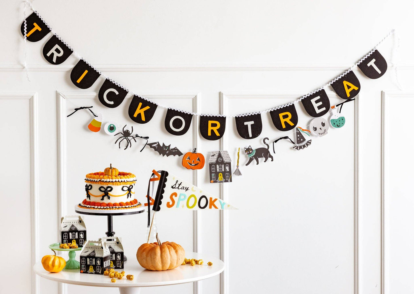 Halloween-themed decor with a 'Trick or Treat' banner, cake, and small houses on a white shelf.